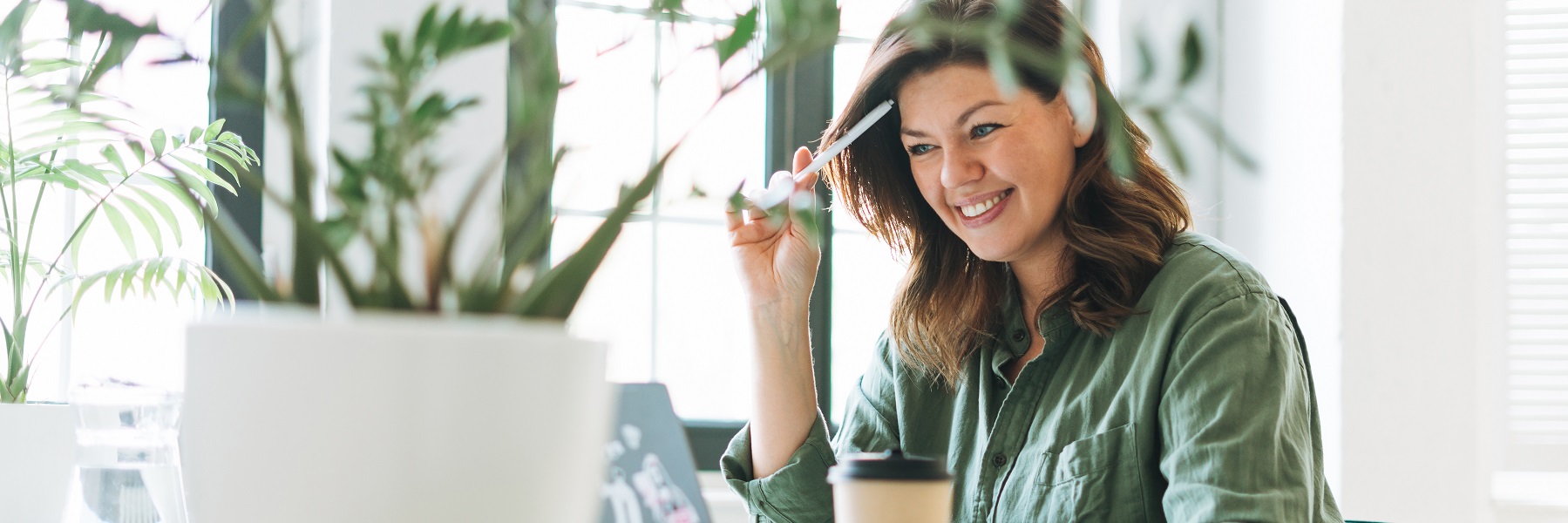 Youngsmilingbrunettewomanplussizeworkingatlaptopontablewithhouseplantinthebrightmodernoffice