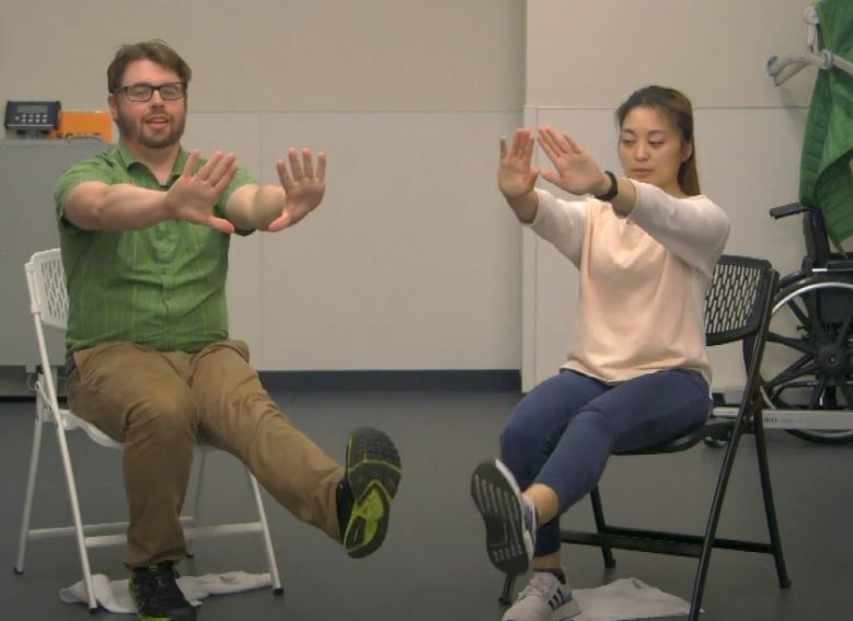 Two physical therapists demonstrate a chair exercise by extending their arms and one leg.
