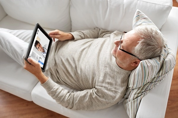 An older man reclines on his couch while video chatting with a doctor.