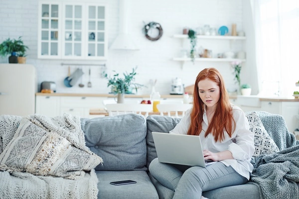 A red-haired woman works on a laptop while sitting on a couch.