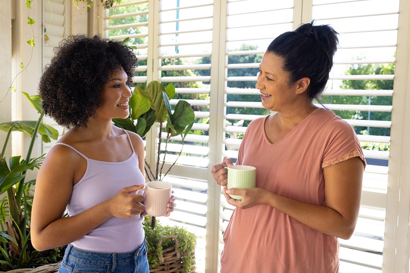 mother and adult daughter holding cups, talking at home