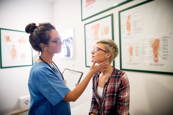 A nurse in blue scrubs examining patient with short hair and glasses