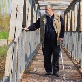 SeniorCaucasianmanwithwalkingstickstandingonanmetalbridgeoversmallriver