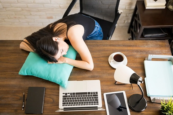 A woman naps at her desk with a laptop and cup of coffee nearby.